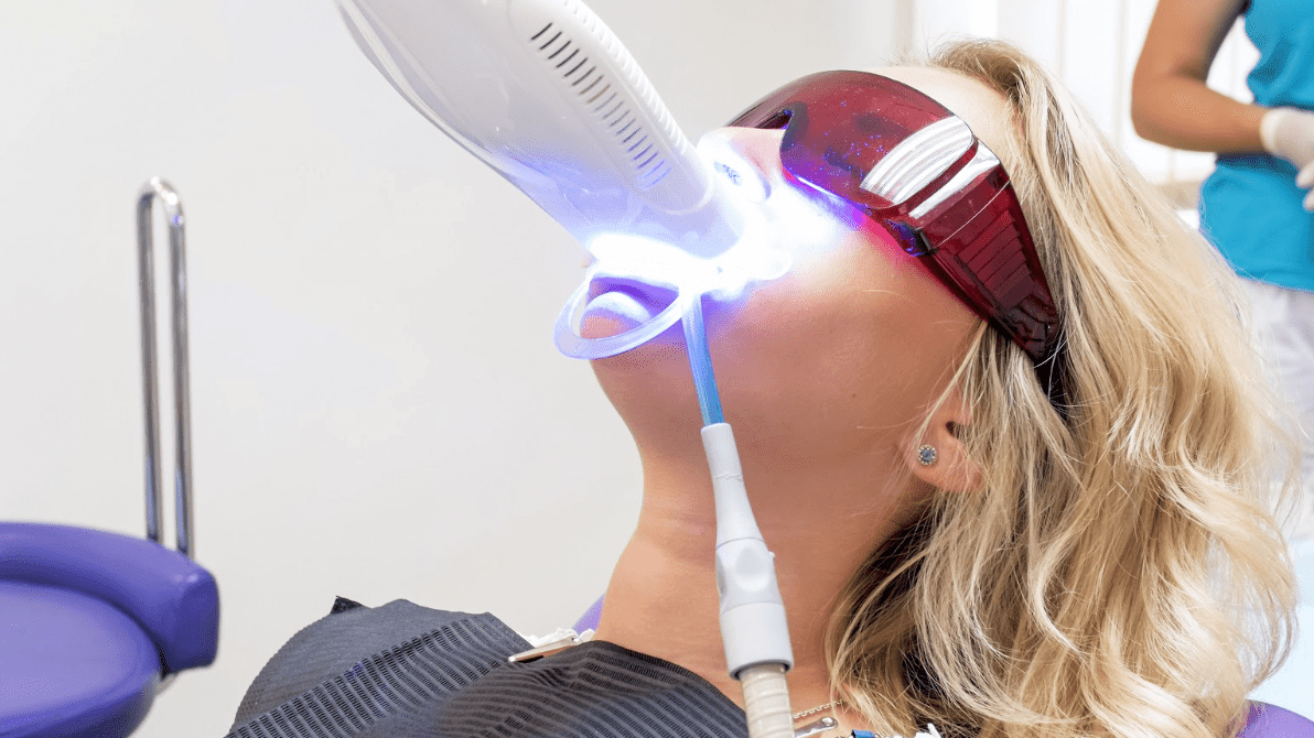 Woman undergoing professional LED teeth whitening treatment at a dental clinic, wearing protective red-tinted glasses while a curing light device emits blue light onto her teeth.