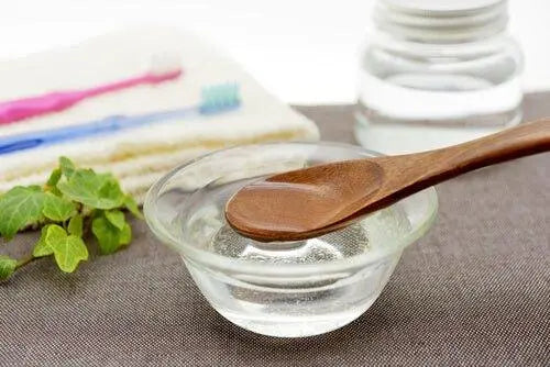 A wooden spoon resting in a glass bowl of coconut oil with toothbrushes and a jar in the background, used for oil pulling oral care.