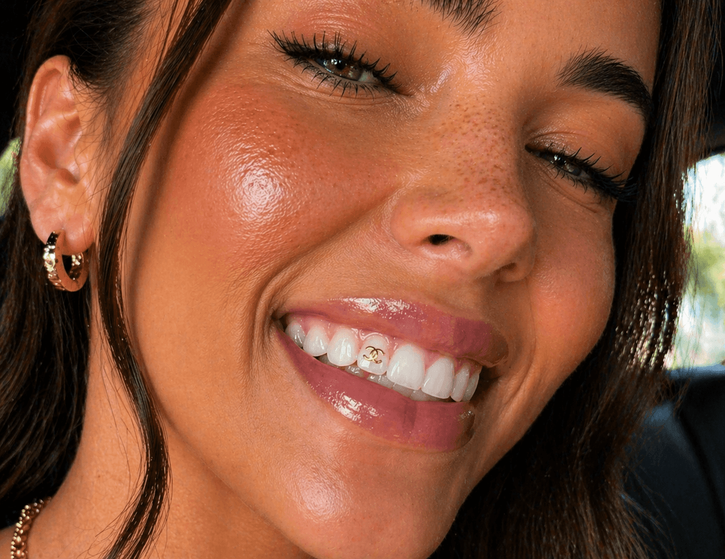 Close-up lifestyle image of a smiling woman winking with a gold Chanel-inspired logo tooth gem on a front tooth, featuring glossy lips and bright white teeth