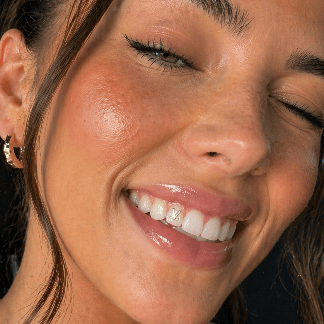Close-up lifestyle image of a woman smiling and winking with a gold LV logo tooth gem on a front tooth, featuring glossy lips and bright white teeth